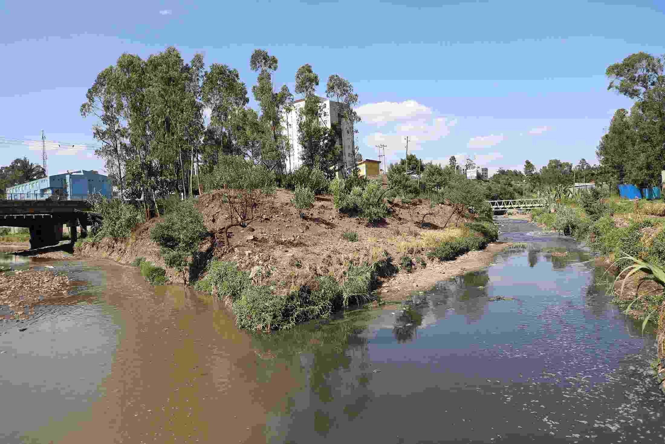 Two parts of a river converge with urban buildings visible behind. The water is murky and polluted. 